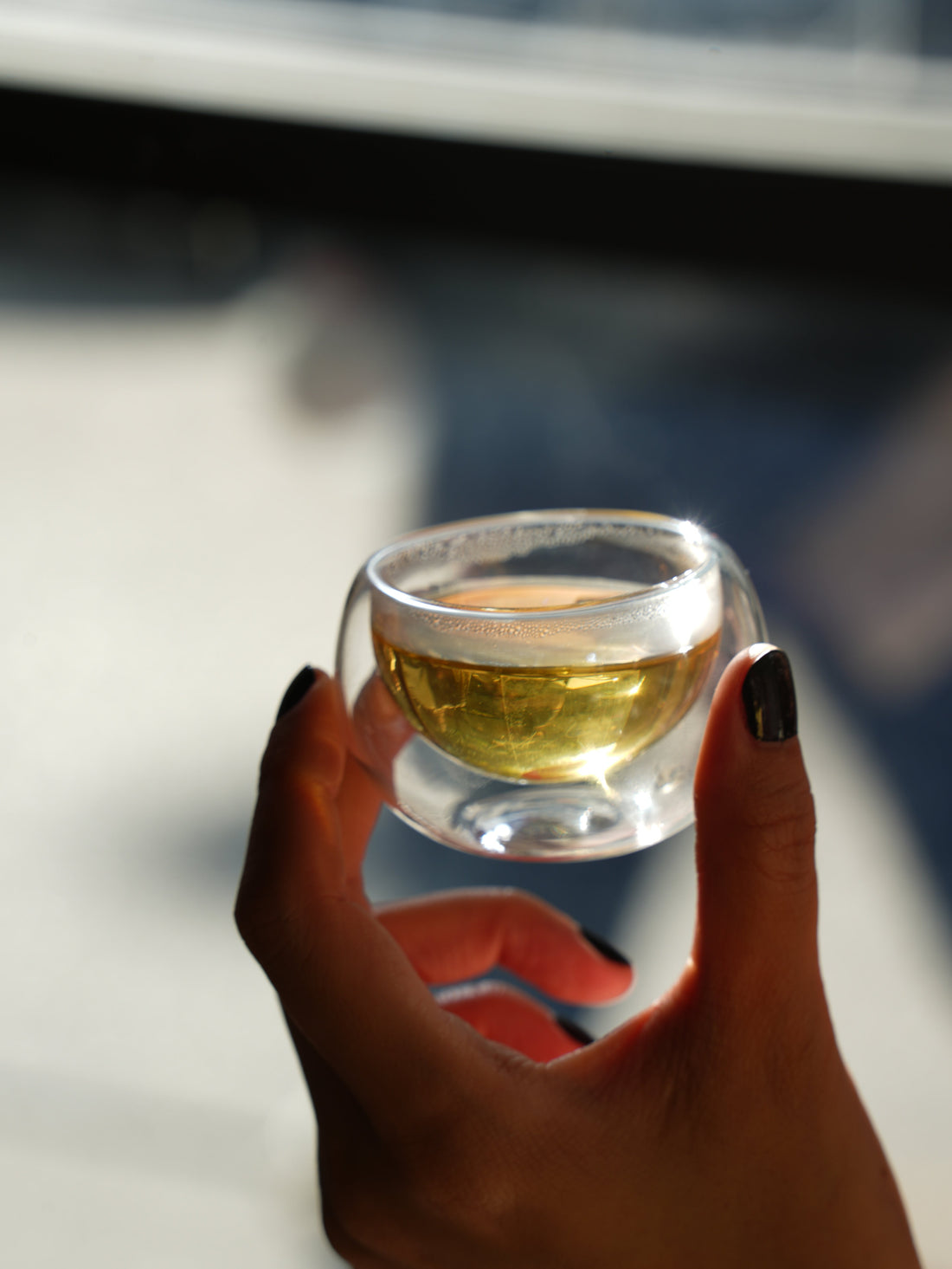 Hand holding a small glass cup of tea in natural light, showing the color and clarity of brewed tea.
