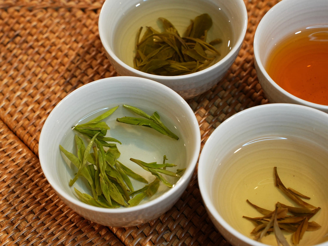 Four porcelain teacups with different loose leaf teas after brewing, showcasing a range of tea colors and infused leaf shapes—highlighting green, yellow, white and black teas on a woven mat background.